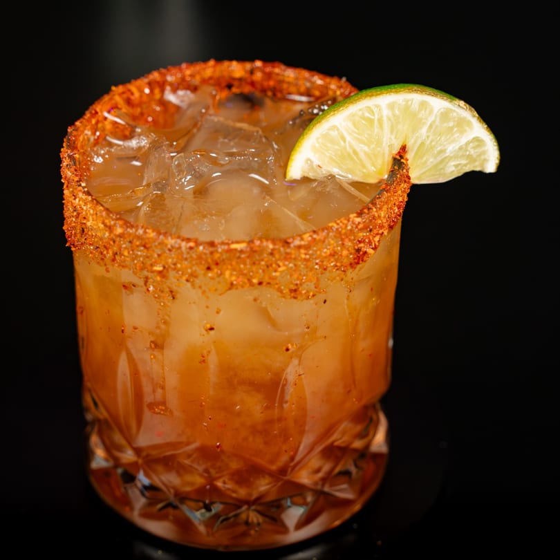 Amber cocktail with ice, a chili-salt rim, and a lime wedge in a faceted glass.