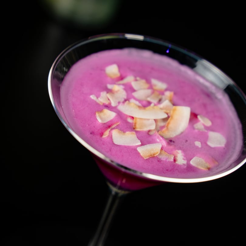 Vibrant pink cocktail in a martini glass, garnished with toasted coconut flakes on a dark background.