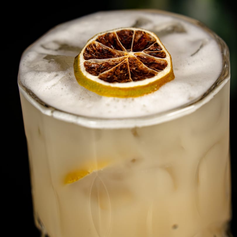 Light-colored cocktail with a thick foamy top, garnished with a dried lime slice.