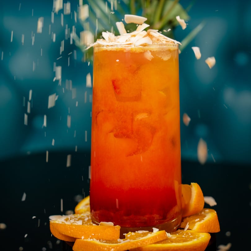 Vibrant orange and red cocktail garnished with falling coconut flakes and fresh orange slices.
