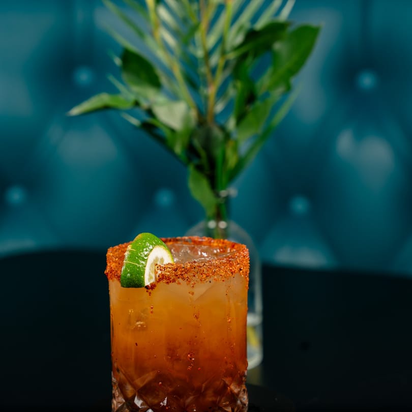 Amber cocktail with a chili-lime rim and lime wedge, set against a blue background.