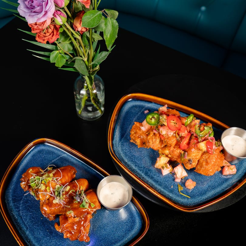 Two blue plates of chicken wings, one with salsa, one with microgreens, beside flowers on a dark table.