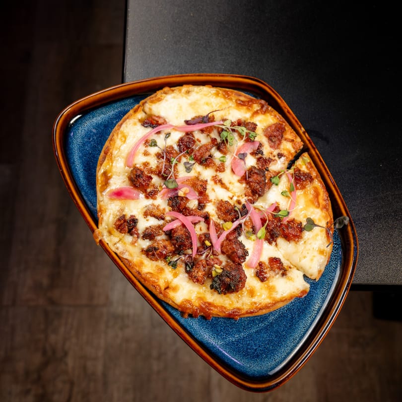 Flatbread pizza with melted cheese, crumbled meat, pickled red onions, and microgreens on a blue plate.